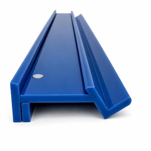 May include: A long, solid blue plastic shelf or ledge with a smooth, glossy finish. The object features a flat horizontal surface, a vertical back panel, and a slanted front edge. A small, circular silver-colored metal fastener is visible on the side.