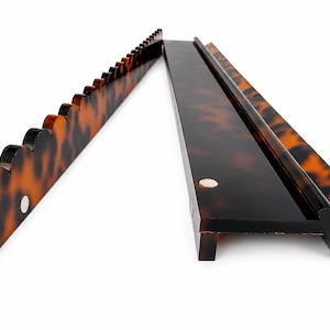 May include: Two long, narrow display racks featuring a brown and black tortoiseshell pattern. One rack has a scalloped top edge with multiple notches, while the other is a flat, L-shaped ledge. Both pieces include small, circular silver-toned magnets.