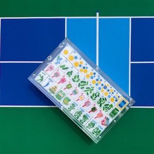 Puede incluir: Un estuche de acr&iacute;lico transparente que contiene un juego de fichas rectangulares con gr&aacute;ficos coloridos. Las fichas presentan ilustraciones de palas de pickleball, pelotas, elementos de pista y la palabra Joker en filas.
