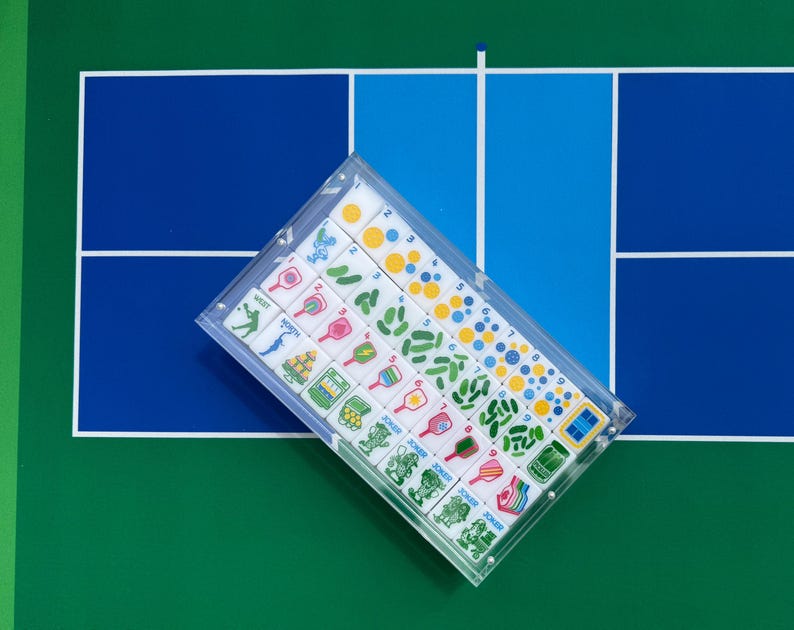 Puede incluir: Un estuche de acr&iacute;lico transparente que contiene un juego de fichas rectangulares con gr&aacute;ficos coloridos. Las fichas presentan ilustraciones de palas de pickleball, pelotas, elementos de pista y la palabra Joker en filas.