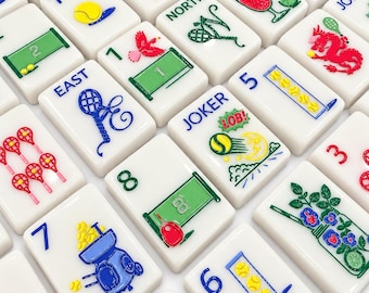 May include: A collection of white rectangular mahjong tiles featuring colorful engraved illustrations related to tennis, including rackets, balls, nets, and court markings. Some tiles display text such as JOKER, POW, NORTH, EAST, and LOB, with various numbers and vibrant graphic designs.