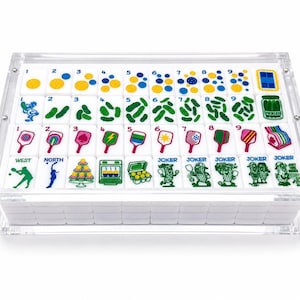 Puede incluir: Un juego de fichas de mahjong blancas con ilustraciones coloridas de paletas de pickleball, pepinillos y n&uacute;meros, alojadas en una caja de acr&iacute;lico transparente. Las fichas presentan varios dise&ntilde;os, incluyendo puntos numerados, pepinillos verdes, paletas rosas y personajes de pepinillos.