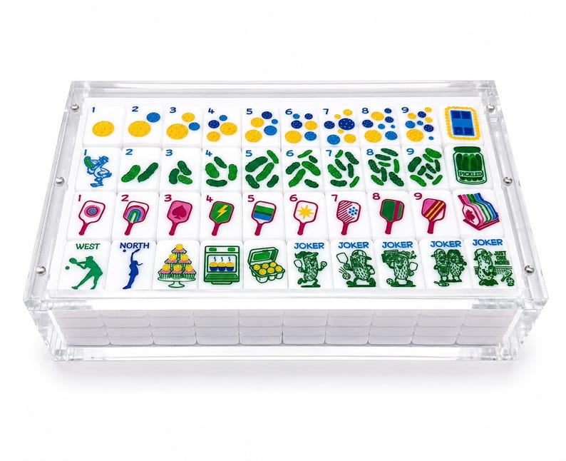 Puede incluir: Un juego de fichas de mahjong blancas con ilustraciones coloridas de paletas de pickleball, pepinillos y n&uacute;meros, alojadas en una caja de acr&iacute;lico transparente. Las fichas presentan varios dise&ntilde;os, incluyendo puntos numerados, pepinillos verdes, paletas rosas y personajes de pepinillos.