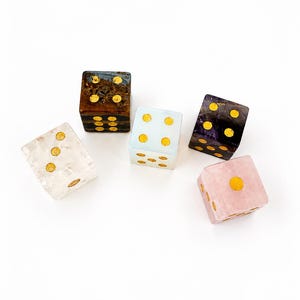May include: A set of five six-sided dice in various colors including translucent white, dark brown, light blue, dark purple, and light pink. Each die features gold-colored circular pips on its faces, arranged in standard numerical patterns.