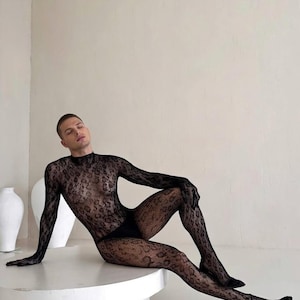 Puede incluir: Un body negro estampado con mangas largas y cuello alto. El body se combina con tacones negros. El atuendo está hecho de un material transparente, similar al encaje. El modelo está sentado en una mesa blanca.