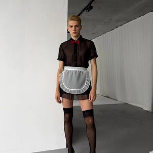Peut inclure: Une chemise noire transparente à manches courtes avec un nœud papillon rouge, un tablier blanc à bordure volantée et une jupe noire. La tenue est complétée par des bas-cuisses noirs et des chaussures à talons hauts.