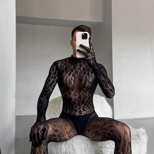 Puede incluir: Un body de encaje negro con estampado, mangas largas y cuello alto. El conjunto incluye guantes y medias a juego. La persona está sentada en una silla blanca y mullida.