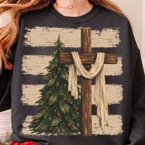 May include: A dark gray crewneck sweatshirt featuring a graphic print of a green Christmas tree decorated with red ornaments next to a brown wooden cross draped with a white cloth, set against a background of horizontal beige brushstroke stripes.