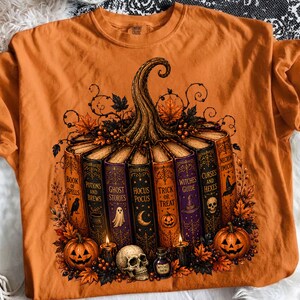 Pode incluir: Uma t-shirt laranja com um gráfico de uma abóbora formada por uma fila de livros com títulos como Book of Spells, Potions and Brews, Ghost Stories, Hocus Pocus, Trick or Treat, Witches Guide, Curses and Hexes e Necronomicon, rodeada de folhas.