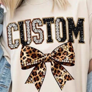 Pu&ograve; includere: T-shirt color crema con la scritta "CUSTOM" in un carattere gioiello. Sotto il testo c'&egrave; un fiocco con stampa leopardata con dettagli dorati e di cristallo. La maglietta &egrave; in stile casual.