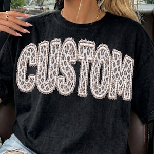 Op de afbeelding: Een zwart t-shirt met een mineral-wash effect en het woord CUSTOM in grote, dikke letters op de borst. De letters hebben een grijs en wit luipaardpatroon met een textuurrijke, gerafelde rand die een gelaagd uiterlijk aan het ontwerp geeft.