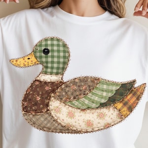 Peut inclure: T-shirt blanc avec un motif de canard en patchwork. Le canard a une tête à carreaux verts, un bec jaune et des ailes faites de divers tissus à motifs dans les tons de marron, vert et crème. Le motif est cousu sur le t-shirt.