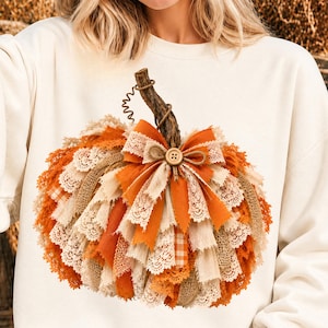 Peut inclure: Un sweat-shirt crème arborant un grand motif de citrouille composé de bandes superposées de tissu orange, de toile de jute et de dentelle blanche. La citrouille possède une tige brune texturée et un nœud central orné d'un petit bouton en bois.