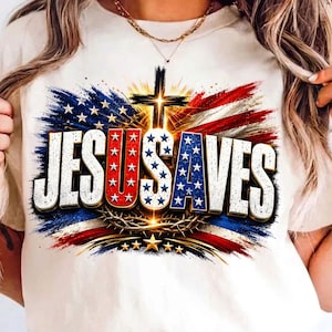 May include: A cream-colored t-shirt featuring a graphic print of a cross and a crown of thorns centered over a stylized American flag. The text "JESUS SAVES" is printed in large, bold letters with a patriotic red, white, and blue star pattern.