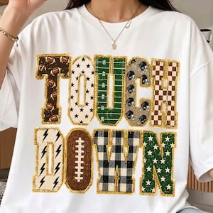 May include: A white short-sleeve t-shirt featuring the word TOUCHDOWN in large block letters. Each letter has a unique pattern including footballs, stars, a green field, helmets, checkers, lightning bolts, a football lace, and plaid.
