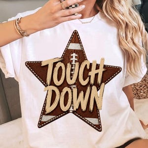 Puede incluir: Una camiseta blanca de manga corta con un gráfico de una estrella marrón grande con textura de balón de fútbol americano. La estrella contiene un diseño de cordones blancos verticales y el texto TOUCH DOWN en letras grandes y desgastadas.