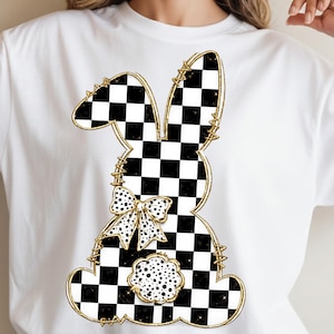 May include: White t-shirt featuring a black and white checkered bunny design with gold accents. The bunny has a bow and a flower-shaped detail. The design is outlined in gold and has a whimsical, Easter-themed aesthetic.