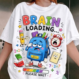 May include: A white t-shirt featuring a colourful graphic with the text BRAIN LOADING... PLEASE WAIT. The design includes a blue backpack character with a sleepy expression, a pencil, a red alarm clock, a maths book, a history book, and a notepad.