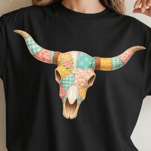 May include: Black t-shirt featuring a colorful, patchwork-style bull skull design. The skull is adorned with various floral and patterned fabrics in shades of pink, blue, yellow, and green. The horns are long and curved, with a brown leather-like band.