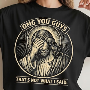 May include: Black t-shirt with a distressed graphic of Jesus covering his eyes. The design includes the text "OMG YOU GUYS" above the image and "THAT'S NOT WHAT I SAID" below.