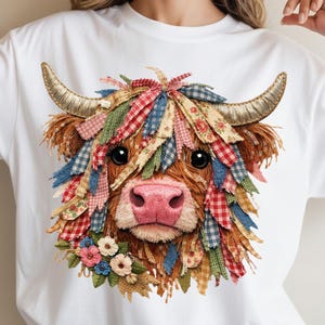May include: White t-shirt featuring a detailed illustration of a Highland cow. The cow has brown fur, a pink nose, and large, dark eyes. Its head is adorned with colorful fabric strips and golden horns. A small floral arrangement is at the bottom.
