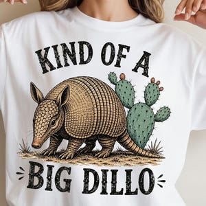 May include: White t-shirt featuring a detailed illustration of an armadillo with a cactus and the text "KIND OF A BIG DILLO" in black. The armadillo is brown and tan, and the cactus is green with orange accents.