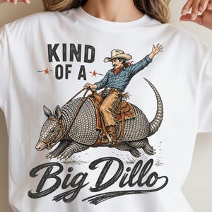 May include: White t-shirt featuring a cowboy riding an armadillo. The cowboy wears a hat, blue shirt, and brown chaps. The armadillo is gray and brown. Text on the shirt reads "KIND OF A" above "Big Dillo".