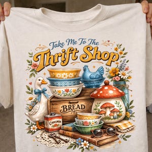 Pode incluir: Camiseta creme com um gráfico colorido que diz "Take Me To The Thrift Shop". O design inclui tigelas vintage, um pato, uma caixa de pão e um recipiente em forma de cogumelo. O design é cercado por flores e outros itens vintage.