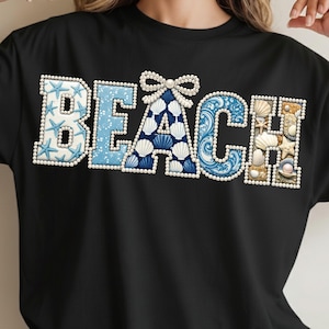 Puede incluir: Camiseta negra con la palabra "BEACH" en un diseño decorativo. Las letras están rellenas de varios elementos con temática de playa, incluyendo estrellas de mar, conchas marinas y un lazo. Las letras están delineadas con pequeñas perlas blancas.