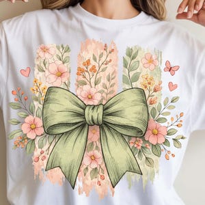 May include: White t-shirt with a green bow and floral design. The design includes pink flowers, green leaves, and a butterfly. The background has vertical stripes in shades of pink and green.