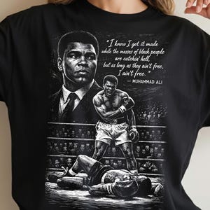 May include: Black t-shirt featuring a black and white graphic of Muhammad Ali. The design includes a portrait, a boxing scene, and a quote: "I know I got it made while the masses of black people are catchin' hell, but as long as they ain't free, I ain't free."