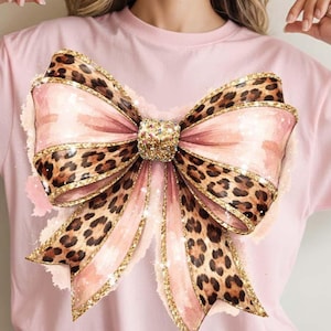 May include: Pink t-shirt featuring a large bow design. The bow has a leopard print pattern with pink and gold accents. The bow's center is embellished with glitter, adding a touch of sparkle to the design. The shirt is a solid pink color.