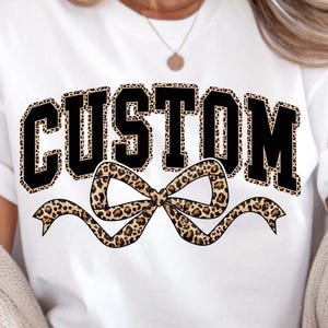 Könnte beinhalten: Weißes T-Shirt mit dem Wort "CUSTOM" in schwarzen Blockbuchstaben mit Leopardenmuster. Unter dem Wort befindet sich eine Schleife mit Leopardenmuster. Das T-Shirt ist ein lässiger Stil.