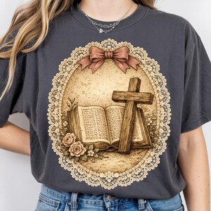 May include: Dark grey t-shirt with a sepia-toned illustration of a wooden cross, open book, and roses inside a lace-edged oval. A brown bow is at the top. The design has a vintage, religious theme.