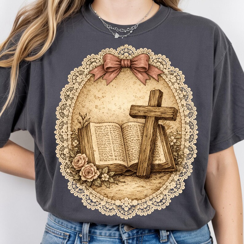 May include: Dark grey t-shirt with a sepia-toned illustration of a wooden cross, open book, and roses inside a lace-edged oval. A brown bow is at the top. The design has a vintage, religious theme.