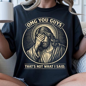 May include: Black t-shirt featuring a distressed graphic of Jesus with his hand over his eyes, encircled by a halo. The text reads "OMG YOU GUYS" and "THAT'S NOT WHAT I SAID". A humorous religious themed tee.