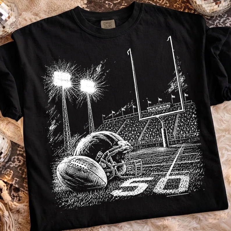 May include: A black t-shirt featuring a white graphic illustration of a football stadium at night. The design includes a football, a helmet, goalposts, stadium lights, and the number 50 printed on the field. The shirt has a crew neckline.
