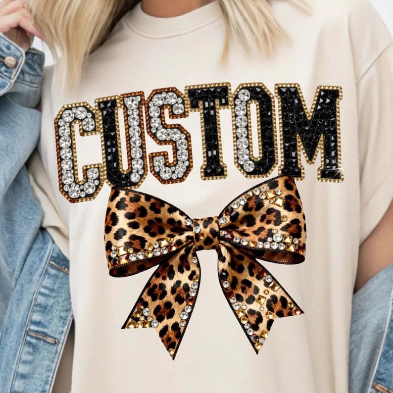 Pu&ograve; includere: T-shirt color crema con la scritta "CUSTOM" in un carattere gioiello. Sotto il testo c'&egrave; un fiocco con stampa leopardata con dettagli dorati e di cristallo. La maglietta &egrave; in stile casual.