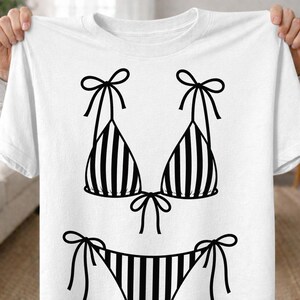 K&ouml;nnte beinhalten: Wei&szlig;es T-Shirt mit einem schwarz-wei&szlig; gestreiften Bikini-Design. Das Bikini-Oberteil hat Schleifen-Tr&auml;ger, und die Unterseite hat Schleifenverschl&uuml;sse an den Seiten. Das Design ist eine einfache, grafische Illustration.