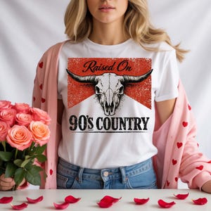 May include: White t-shirt featuring a bull skull graphic with the text "Raised On 90's Country" in black. The design has a distressed, vintage look with a red background. The shirt is a classic crew neck style.