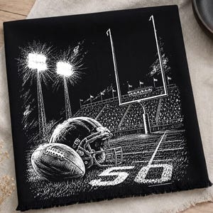 May include: A black fabric square featuring a white screen-printed illustration of a football stadium at night. The design includes a football, a helmet, goalposts, stadium lights, and the number 50 printed on the field with a fringed edge.