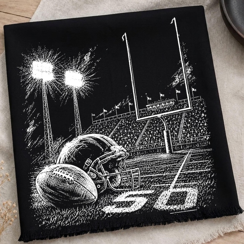 May include: A black fabric square featuring a white screen-printed illustration of a football stadium at night. The design includes a football, a helmet, goalposts, stadium lights, and the number 50 printed on the field with a fringed edge.