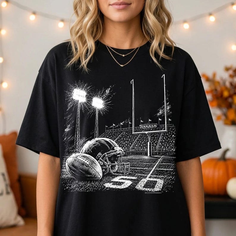 May include: A black t-shirt featuring a white graphic print of a football stadium at night. The design includes a football, a helmet, goalposts, stadium lights, and the number 50 on the field, rendered in a detailed, sketch-like artistic style.