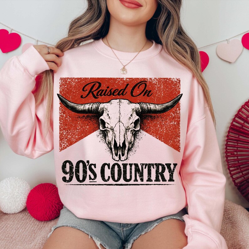 May include: A light pink sweatshirt featuring a graphic of a bull skull with the text "Raised On 90's Country" in black. The design is set against a red and white background.