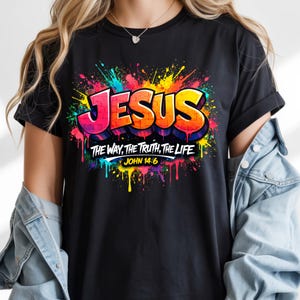 May include: Black t-shirt with a colorful graphic design. The design features the word "JESUS" in bold, outlined letters, with the text "THE WAY, THE TRUTH, THE LIFE" and "JOHN 14:6" below. The design has a graffiti-style aesthetic with paint splatters.