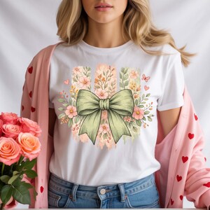 May include: White t-shirt with a large green bow design, floral accents, and vertical pastel stripes. The design incorporates pink flowers, butterflies, and leaves, evoking a spring or summer aesthetic.