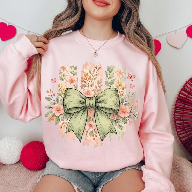 May include: A pale pink sweatshirt with a large green bow and floral design. The design includes pink flowers, butterflies, and vertical stripes with a watercolour effect. The sweatshirt is a crew neck style.