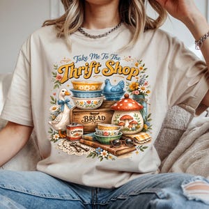 K&ouml;nnte beinhalten: Beiges T-Shirt mit einem farbenfrohen Aufdruck mit der Aufschrift "Take Me To The Thrift Shop". Die Grafik zeigt Vintage-Artikel, darunter Schalen, eine Teekanne, eine Gans und Blumen. Das Wort "Bread" steht auf einer Holzkiste.