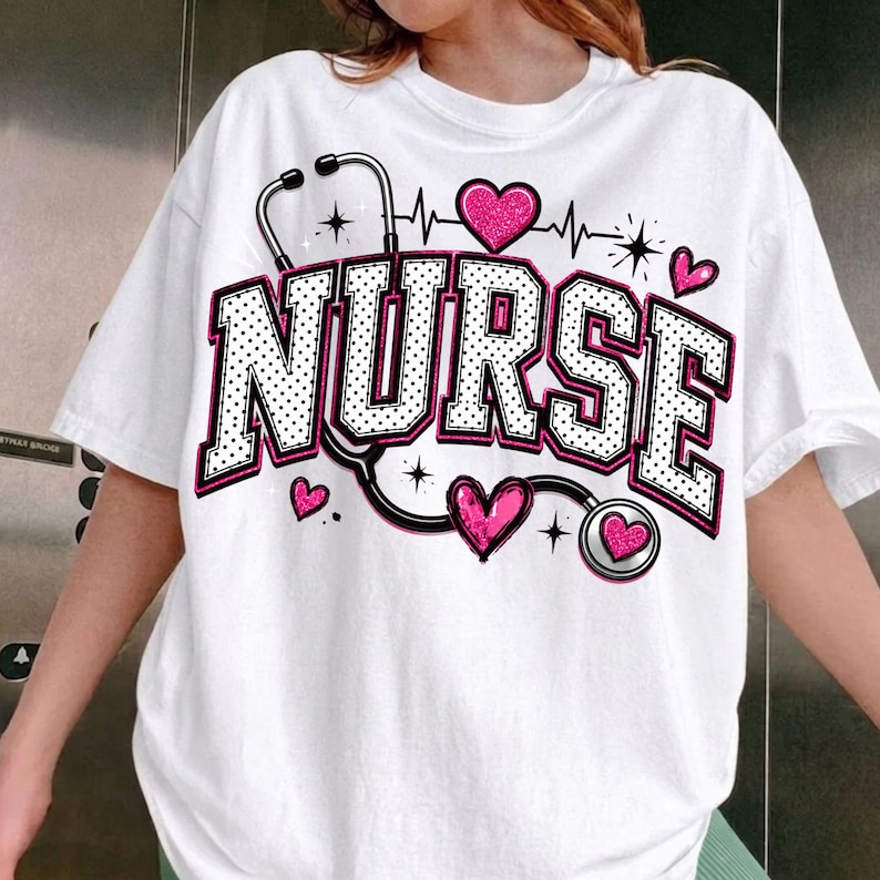 Puede incluir: Una camiseta blanca de manga corta con la palabra NURSE en letras grandes de bloque con lunares blancos y negros y borde rosa. Un gr&aacute;fico de estetoscopio negro rodea el texto, acentuado por varios corazones rosas brillantes.