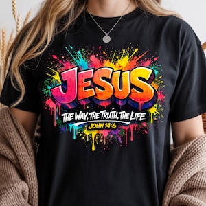 May include: Black t-shirt featuring the word "JESUS" in vibrant graffiti-style lettering, with a colorful paint splatter background. The text "THE WAY, THE TRUTH, THE LIFE" and "JOHN 14:6" are also displayed. A casual, graphic tee.